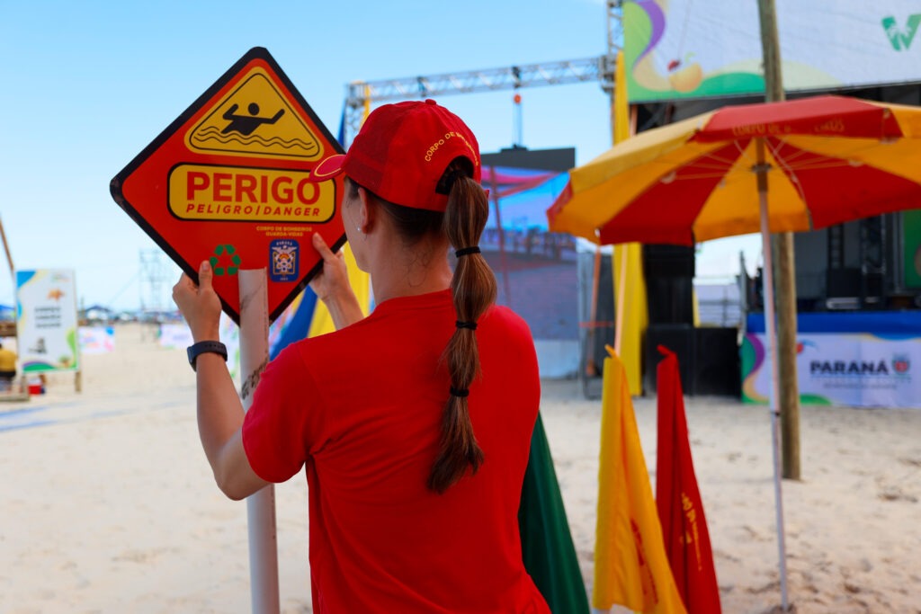 Pessoa de costas para a câmera manipula placa de metal. A pessoa tem cabelo longo e está vestindo camiseta e boné vermelhos. A placa de metal, virada para a câmera, diz: "Perigo, peligro, danger"