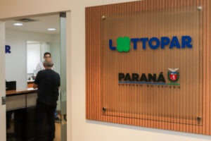 A imagem mostra uma placa de vidro com a inscrição "Lottopar" em letras azuis, com o segundo "o" substituído por um trevo verde de quatro folhas, simbolizando sorte. Abaixo, está o texto "Paraná - Governo do Estado", com o brasão do Paraná à direita. A placa está fixada em uma parede revestida com ripas de madeira. Ao lado, é possível ver o interior de um escritório, com uma pessoa de costas, aparentemente em atendimento, e outra ao fundo, vestida com uma camisa branca.