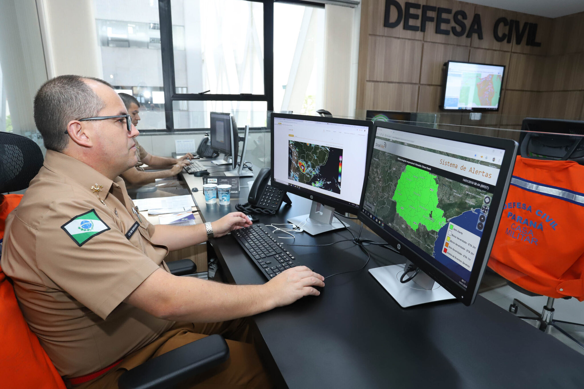 Pessoa fardada utiliza computador com dois monitores. Em um deles, o mapa do estado do Paraná está preenchido na cor verde neon