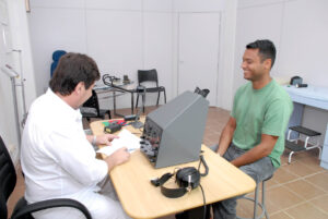 A imagem retrata uma consulta em um ambiente clínico. Um homem de cabelos curtos, vestido com uma camiseta verde, está sentado sorrindo em frente a uma mesa. Do outro lado da mesa, um profissional de jaleco branco está lendo e preenchendo documentos. Na mesa, há um equipamento eletrônico com botões e mostradores, além de um par de fones de ouvido, sugerindo um exame técnico, possivelmente relacionado à audição ou reflexos.