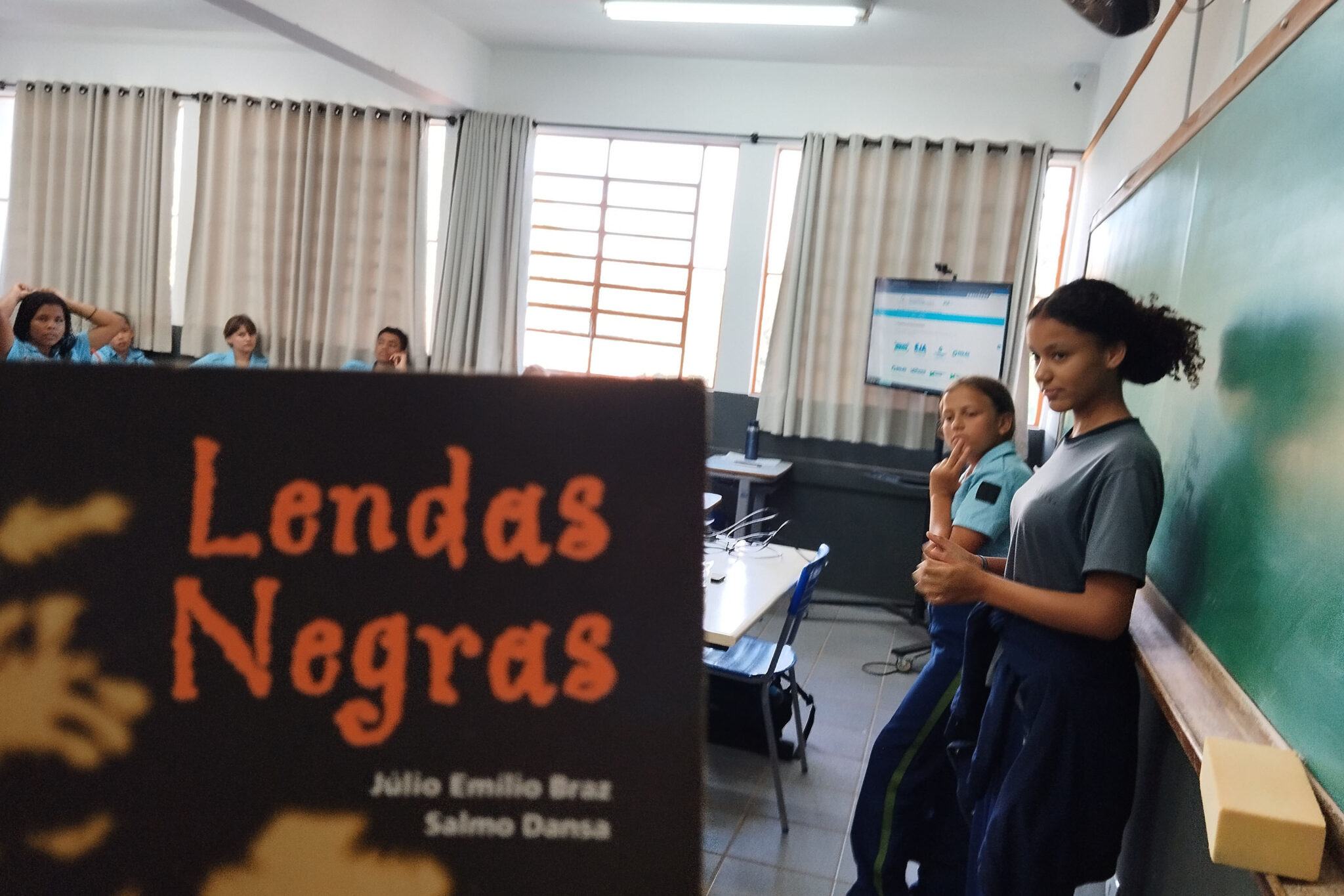 A imagem mostra uma sala de aula com cortinas abertas e janelas que permitem a entrada de luz natural. Em primeiro plano, há a capa do livro "Lendas Negras", de Júlio Emílio Braz e Salmo Dansa, desfocada, destacando o título em laranja sobre um fundo preto. Ao fundo, duas alunas estão de pé próximas ao quadro verde, uma delas gesticulando e a outra observando, ambas vestindo uniformes escolares. Mais ao fundo, outros estudantes sentados em suas carteiras participam da atividade.