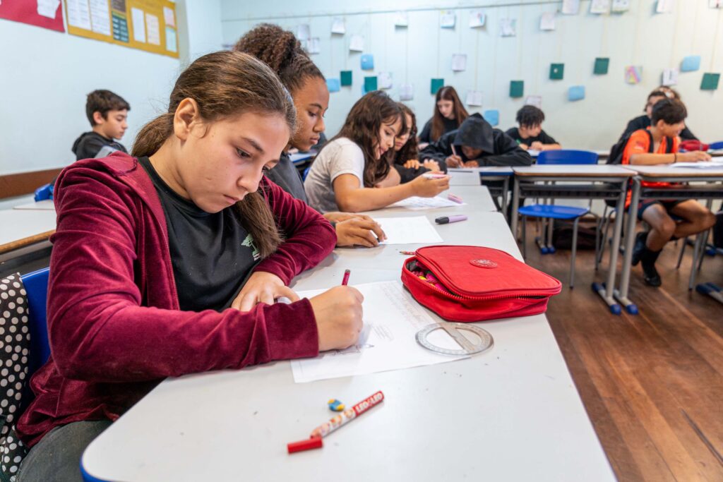 Sala de aula com mesas organizadas em forma de ferradura. Em primeiro plano, uma criança escreve em um papel