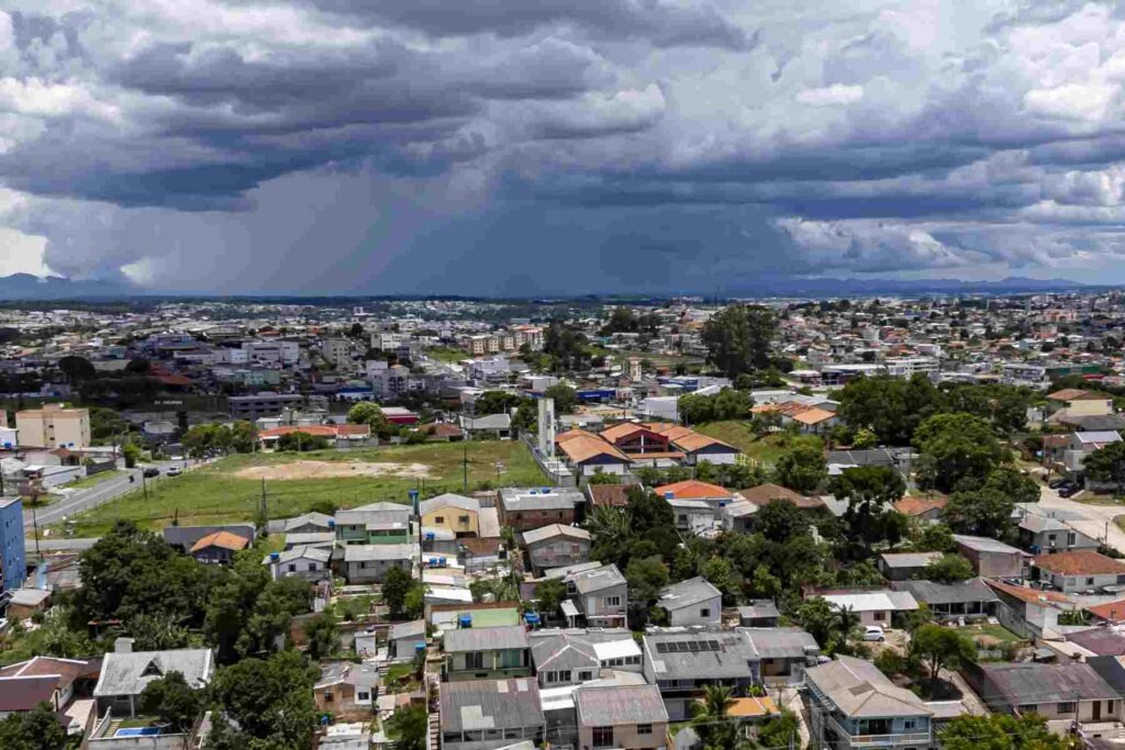Imagem áerea mostra casas e prédios de uma cidade com nuvens carregadas de chuva ao fundo