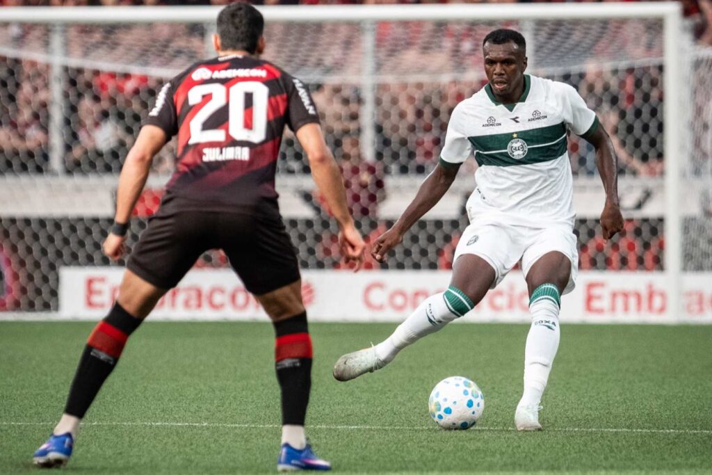 O jogador Jacy, do Coritiba, passa a bola com a perna direita sendo observado de frente pelo atleta Julimar, do Athletico